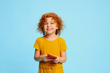 Portrait of cute little girl, child with curly red hair playing on phone, smiling isolated over...
