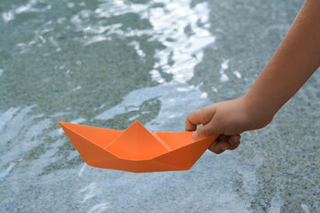 Kid launching small orange paper boat on water outdoors, closeup