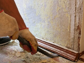 a builder breaks out a wooden plinth in an old house in the process of a major renovation, working with a crowbar to remove a floor wall panel in a room with crumbling plaster and construction dust
