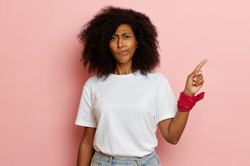 Young curly haired female stands isolated over pink wall and shows finger aside, wearing white t-shirt and red bandana on the arm