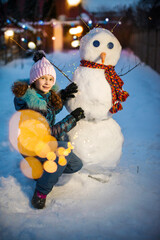 Happy girl playing with snowman on snowy winter walk in evening near the house. Outdoor fun for...