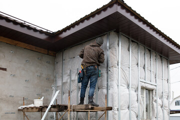 Naklejka na ściany i meble A man installs metal supports on the walls of the house for siding