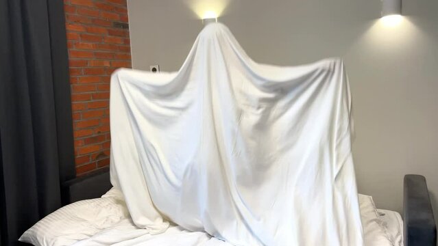 kid playing ghost inside blanket sheet in bedroom. boy jumping having fun halloween holiday crazy