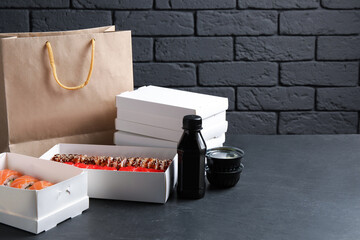 Food delivery. Boxes with delicious sushi rolls, paper package and soy sauce on black table, space...