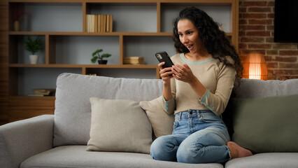 30s Hispanic Latina Caucasian girl woman lady sitting on couch at home living room play with mobile phone win game online betting victory has discount in net virtual hop e-commerce triumph celebrate