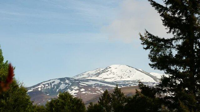 Time lapse of clouds swirling past snow capped hills behind trees on a sunny afternoon