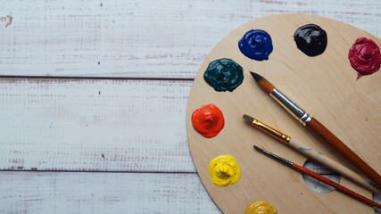 Artist's palette with samples of colorful paints and brushes on white wooden table, top view. Space...
