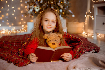 a little girl reads a book with a teddy bear near the Christmas tree at home for Christmas