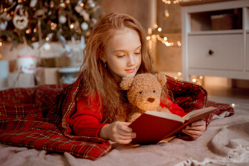 a little girl reads a book with a teddy bear near the Christmas tree at home for Christmas