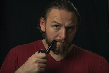Lumberjack brutal beard muscled man in red shirt with smoking tube pipe standing on dark background