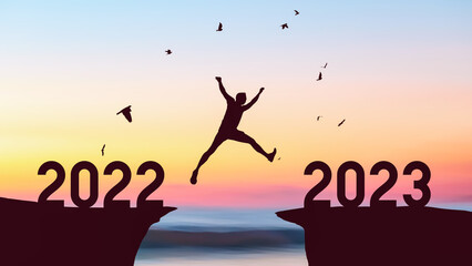 Silhouette man jumping between cliff with number 2022 to 2023 and birds flying at tropical sunset...