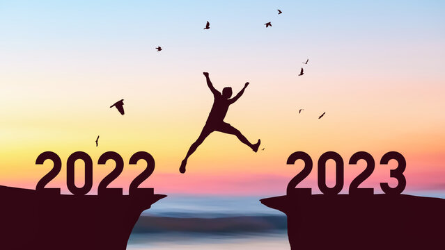 Silhouette man jumping between cliff with number 2022 to 2023 and birds flying at tropical sunset beach. Freedom challenge and travel adventure holiday concept.