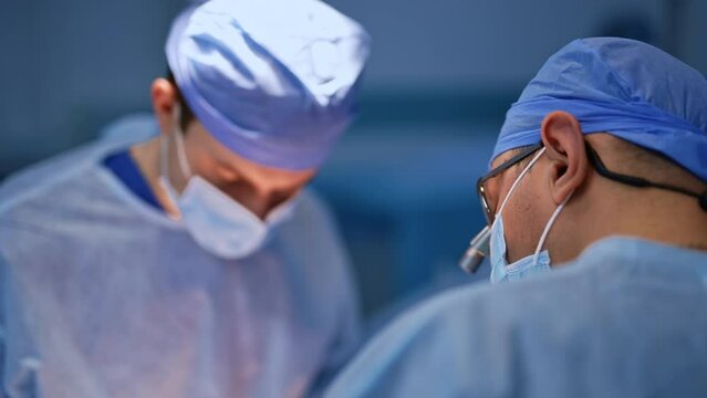 The doctor in device glasses rear view at operation. Close up. Younger assisting surgeon stands his face to camera looking down. Blurred backdrop.