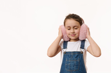 Adorable Caucasian child, fashionable cute baby girl wearing denim overalls, warming her ears with her new fashionable pink fluffy clothing accessory - plush earmuffs. Copy space on white background