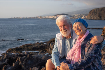 Fototapeta premium Smiling beautiful senior couple sitting on the rocks at sea enjoying travel and nature at sunset light. Relaxed lifestyle for a caucasian couple of retirees