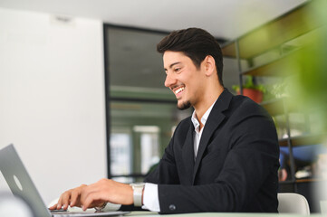 Happy smart young hispanic businessman wearing formal suit using the laptop computer at modern office. Smiling latin male office employee at the workplace is typing