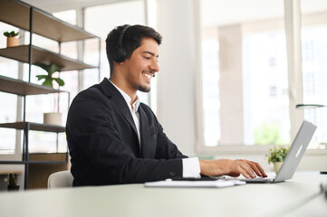 Young man wearing wireless headset using a laptop computer for communication with customers or colleagues sitting at the desk in contemporary office. Maale support worker talks online. Side view