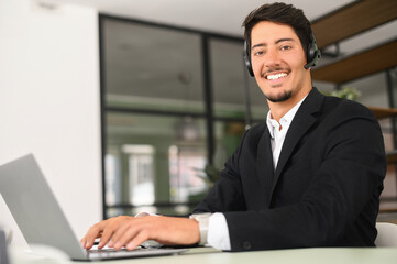 Fototapeta na wymiar Friendly and smiling hispanic man wearing wireless headset looks at the camera sitting at the desk in front of laptop in modern office, male employee using hands free device for online connection
