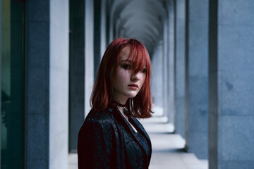 Young woman wearing long black cloak with red hair standing in corridor between the columns of architectural building