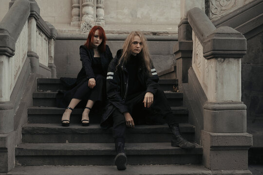 Young man and woman couple gothic style sitting on the steps of the gothic cathedral of architectural building. A guy wearing long blond hair and a girl with red hair