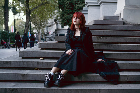 Young woman dressed gothic style in long black dress and cloak wearing red hair sitting sad and pensive outdoors in city on the steps of medieval European building