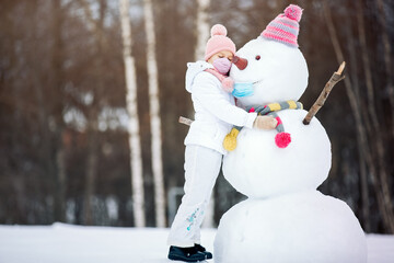 A child girl wearing a mask herself and on a snowman ,playing with a snowman on a winter walk in...