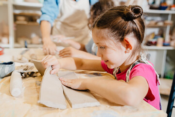 Kids sculpting clay crafts while sitting at table in pottery class - obrazy, fototapety, plakaty