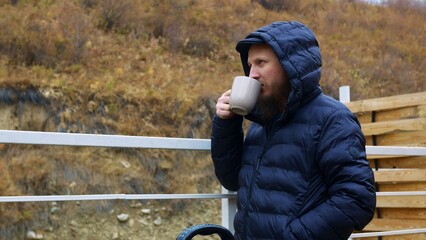 Fototapeta premium Bearded millennial man stands by a charcoal grill and enjoys his mug of coffee while waiting for food to be prepared to eat in nature. A man in a mountainous area drinks coffee and admires the scenery
