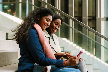 Two young cheerful multinational women using cellphone while sitting on stairs