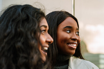 Two young multinational women looking aside while standing at the city street