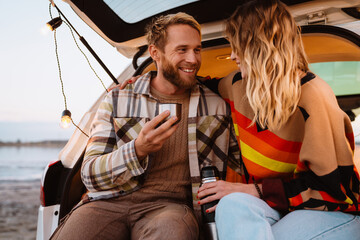 Happy young white couple drinking tea while sitting in trunk by seaside