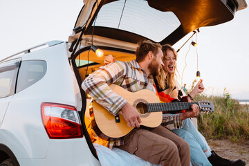 Happy young couple playing guitar while sitting in trunk by seaside