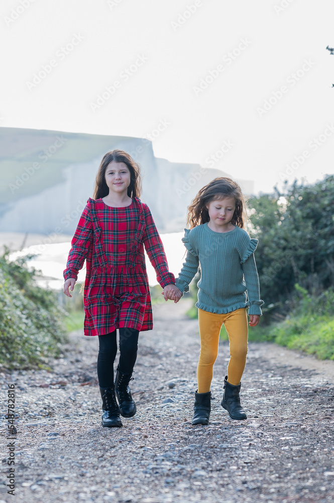 Wall mural two little girls in red checked dress and yellow trousers walking holding hands back from cuckmere b