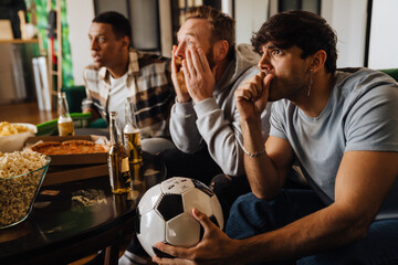 Upset male friends watching sport game and drinking beer at hone