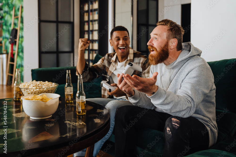 Wall mural Two excited men playing video games and drinking beer in living room