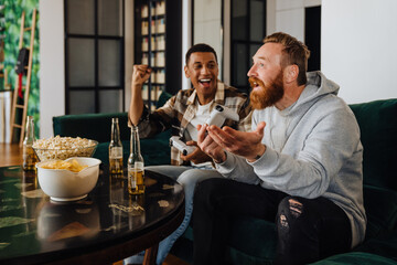 Two excited men playing video games and drinking beer in living room