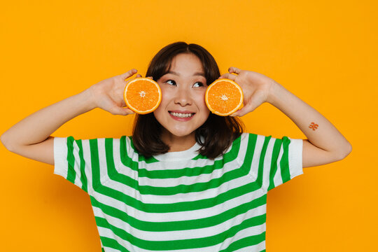 Cheerful asian girl holding slices of orange in her hands isolated over yellow background