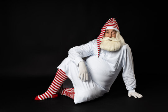 Santa Claus in pajamas sits on his side in the dark and looks straight into the camera. The concept of an unusual Santa on Christmas night. Photo on a black background.