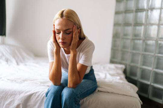 Portrait of worried millennial blonde female holding head in hands feeling anxiety sitting in living room at home. Frustrated depressed young woman suffering from emotional stress sitting alone on bed