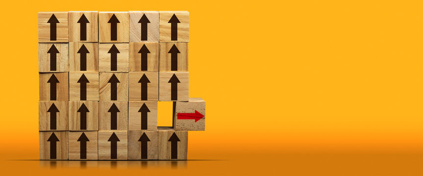 Concept of standing out from the crowd. Group of wooden blocks with brown arrows and a wooden cube with red arrow moving away, on a yellow and orange background with copy space and reflections.