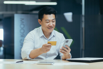 Young Asian man sitting at desk in office, paying with credit card online, making order by phone, making transaction with mobile banking app.