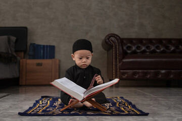 Fototapeta na wymiar Cute little Muslim boy wearing traditional cloth holds rosary in hand, sit on praying mat and interested in the Quran the holy book of Muslims