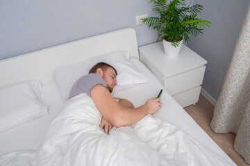 A man with a beard uses phone in his home bed. Shopping for goods without getting out of bed. The concept of people's dependence on gadgets. Internet addiction. Dependence on phone. Gadget addiction.