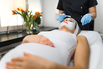 young Caucasian pregnant woman lying on a table and a cosmetologist making a facial mask for her,...
