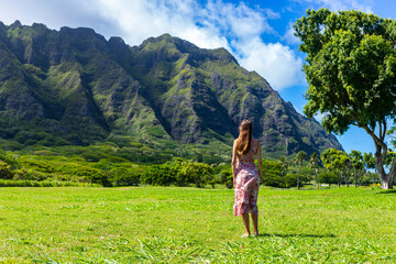 girl in long dress walks on grass at kualoa point on oahu, hawaii, overlooking mighty green...