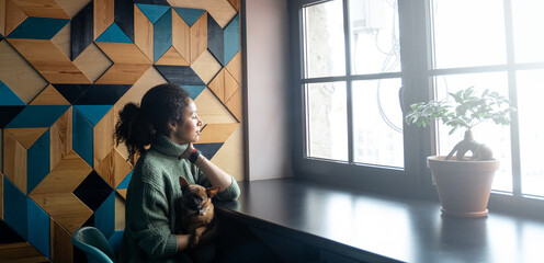 Beautiful black woman with pet dog sitting in cafe waiting for coffee, looking at window and...