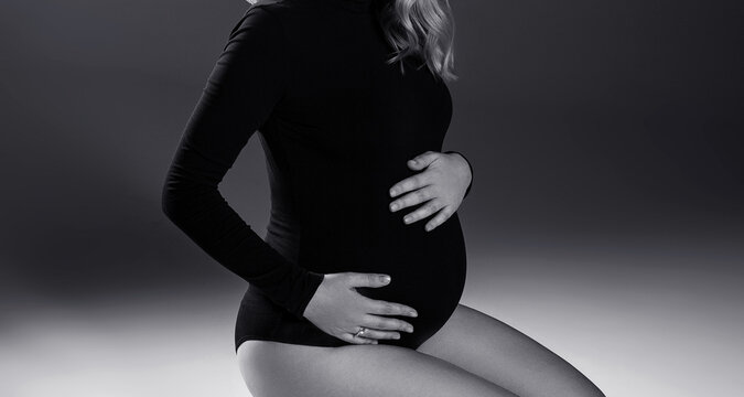 A pregnant woman in a black bodysuit sits on a gray background. Pregnancy. Studio shooting. Black and white photo. Woman in black.
