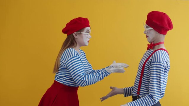 A couple of young mimes, a girl and a guy, dressed in bright clothes and make-up arguing with each other, actively gesticulating, waving their arms, on a yellow background in studio