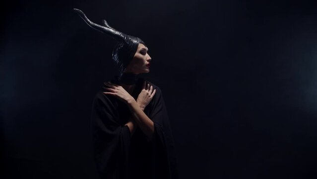Beautiful woman in a dark costume. A woman seduces with hand gestures and a mystical look. A girl in a black devilish costume with horns on a black background. 