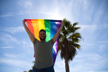 Handsome young gay man holding the gay pride flag in the wind, blue sky in the background. The flag...